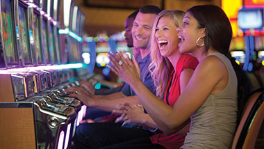 group of people sitting at a row of slot machines playing loughing and clapping hands